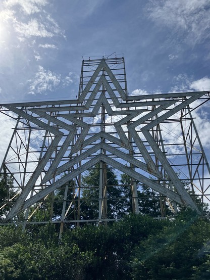 A large, weathered metal star structure stands against a blue sky with clouds, partially obscured by trees. At night, the star is lit in red and white neon lights and can be seen all throughout the valley. 