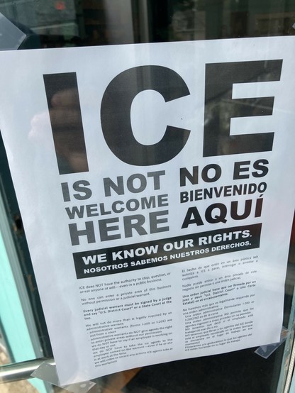 A printed sign displayed on a door, stating "ICE IS NOT WELCOME HERE" in bold letters. The sign includes a message in both English and Spanish explaining rights related to Immigration and Customs Enforcement (ICE), emphasizing that ICE does not have the authority to enter without a warrant. 