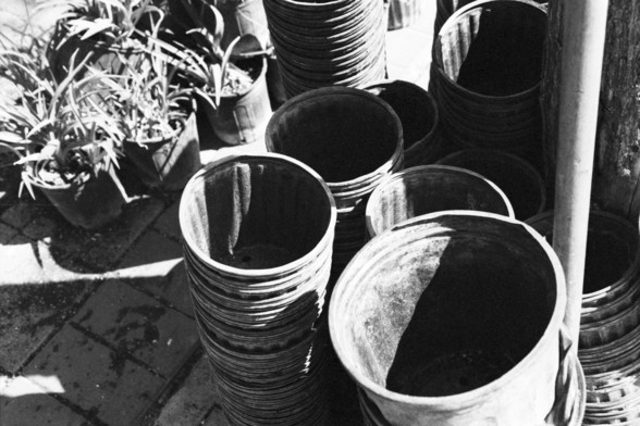 **English:**
This black-and-white photograph captures a collection of empty plant pots arranged in a somewhat haphazard manner. The pots vary in size and are stacked or placed next to each other. Some pots are upright, while others are slightly tilted. In the background, there are a few potted plants with visible foliage, adding a touch of greenery to the scene. The ground appears to be paved with bricks, and the lighting suggests it might be outdoors, possibly in a garden or nursery setting.

…