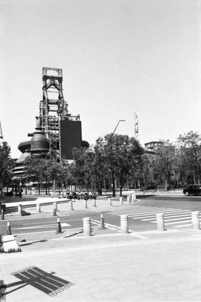 **English:** A black and white photograph of an industrial area with a large structure in the background, possibly a blast furnace or industrial tower. The foreground features a pedestrian crossing and some trees.

**Chinese:** 这张黑白照片展示了一个工业区，背景中有一个大型结构，可能是一个高炉或工业塔。前景是一个行人过街处和一些树木。