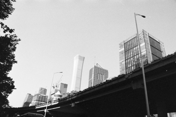### Image 3:
**English:** A black and white photograph of a cityscape with modern skyscrapers in the background. The foreground features an elevated road or bridge with street lamps.

**Chinese:** 这张黑白照片展示了一个城市景观，背景中有现代化的摩天大楼。前景是一条高架路或桥梁，配有路灯。