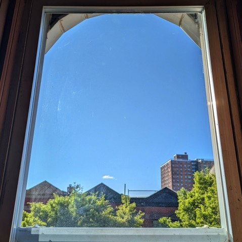 Looking through an arched window two hours after sunrise the sky is bright blue, with one tiny puffy white cloud just above the horizon. Pointed roofs of Harlem brownstones are silhouetted across the street, and a taller apartment building can be seen in the distance. The green tops of two trees are on the bottom and right. 