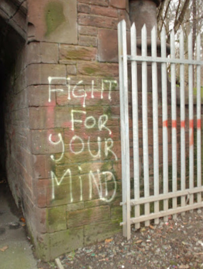Photo of a corner of a dusty old brick wall, with a metal white picket fence leaning against it on the right. Painted on the wall, in thin white letters, “Fight for your mind”
