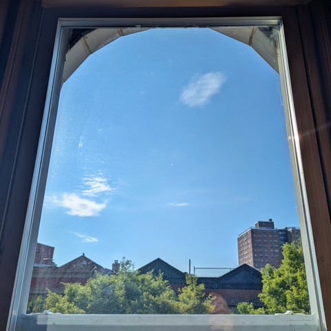 Looking through an arched window two hours after sunrise the sky is bright blue with a few scattered white clouds. Pointed roofs of Harlem brownstones are silhouetted across the street, and a taller apartment building can be seen in the distance. The green tops of two trees are on the bottom and right.