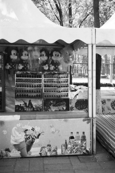 **English:**
This black-and-white photograph depicts a small outdoor kiosk or stall. The kiosk is adorned with various plush toys, prominently featuring several Mickey Mouse dolls hanging at the top. Below the toys, there are shelves stocked with small items, possibly keychains or small trinkets. The front of the kiosk displays images of ice cream cones and bottles of beverages, suggesting that it might also sell refreshments. The setting appears to be a park or a fair, with trees and a fence v…