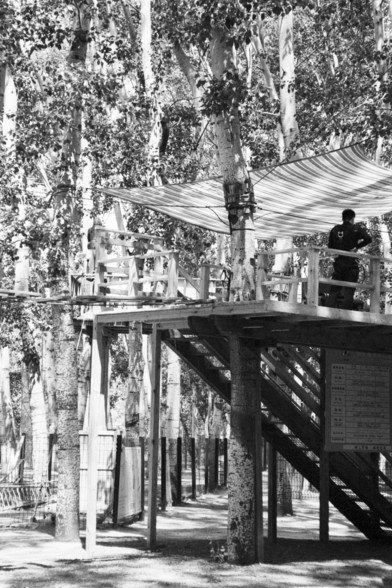 English alt text: A wooden platform nestled among tall trees, covered by a striped canopy. A person stands on the platform, which is accessed by a stairway. Beneath the structure is a bilingual sign, possibly in Chinese, and fencing and foliage frame the background, suggesting a serene park or nature retreat.

中文替代文本（Chinese alt text）：一座建在高大树木之间的木质平台，上方覆盖着条纹遮阳篷。一人站在平台上，通往平台的楼梯清晰可见。平台下方有一块双语标牌（可能含有中文），背景中的树木和围栏营造出宁静的自然氛围。