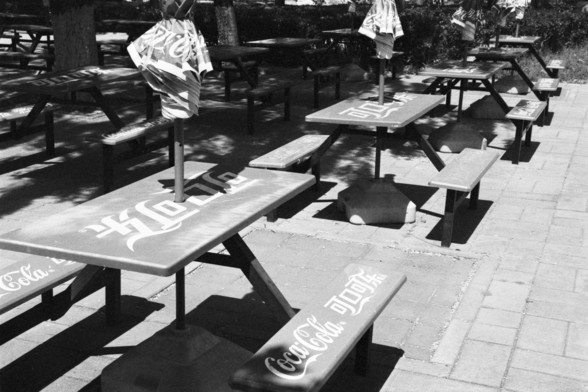 **English:**
This scan from a black and white film negative shows an outdoor seating area, likely part of a park or recreational facility. The seating consists of several picnic tables, each adorned with Coca-Cola branding. The tables are arranged neatly on a paved surface, and each table is equipped with an umbrella providing shade. The area is surrounded by trees, creating a pleasant and shaded environment. The overall scene suggests a place where people can relax and enjoy a meal or a drink …