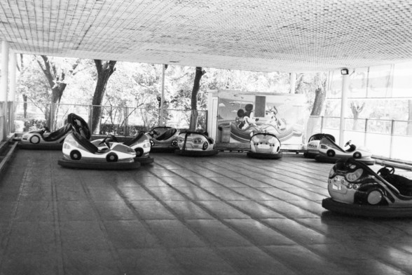 **English:**
This scan from a black and white film negative depicts an indoor bumper car arena. The scene is devoid of people, giving it a quiet and somewhat nostalgic atmosphere. Several bumper cars are scattered across the floor, each equipped with large rubber bumpers. The background features large windows that allow natural light to filter in, illuminating the space and casting shadows. Outside the windows, trees can be seen, suggesting that the arena is located in a park or a similar outdo…
