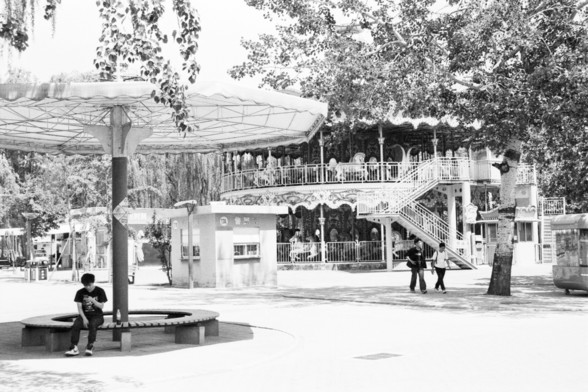 **English:**
This scan from a black and white film negative features a classic carousel situated in an outdoor setting, likely within an amusement park. The carousel is a two-tiered structure with ornate decorations and a canopy roof. Several people can be seen enjoying the ride, adding a sense of movement and joy to the scene. In the foreground, a person is seated on a bench under a large umbrella, while another person is walking nearby. The area is surrounded by trees, providing a natural bac…