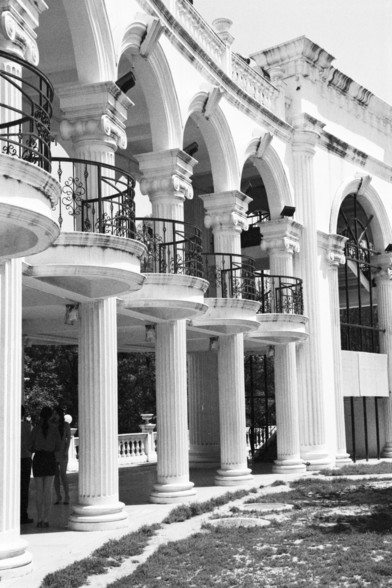 **English:**
This black and white image showcases a grand architectural structure adorned with classical elements. The building features a series of ornate arches supported by intricately designed columns. Balconies with wrought iron railings overlook the scene, and the structure is embellished with decorative moldings and carvings. People can be seen standing under the arches, providing a sense of scale to the imposing architecture.

**Chinese:**
这张黑白图片展示了一座装饰有古典元素的宏伟建筑结构。该建筑具有精心设计的柱子支撑的一系列装饰性…