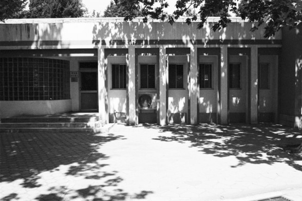 **English:**
This black and white photograph depicts the exterior of a modern building with a minimalist architectural design. The building features a flat roof and a series of evenly spaced vertical columns that create a rhythmic pattern of light and shadow. The facade includes several narrow windows, and there is a small set of stairs leading up to a door on the left side. The shadows of trees are cast onto the building, adding a natural element to the otherwise geometric scene.

**Chinese:**…