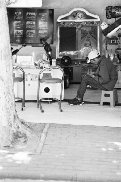 **English:**
A black-and-white photograph depicting a man sitting on a small stool outside a small amusement arcade. The man is wearing a cap and appears to be looking at his phone. The arcade features various game machines and a large crane game machine prominently displayed. There are several empty chairs in front of the arcade, and a tree is visible on the left side of the image.

**Chinese:**
一张黑白照片，描绘了一名男子坐在游乐场外的小凳子上。该男子戴着帽子，似乎正在看手机。游乐场内有各种游戏机，其中一个大型抓娃娃机非常显眼。游乐场前有几把空椅子，照片左侧可以看到一棵树。
