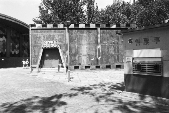 **English:**
A black-and-white photograph showing the entrance to an attraction labeled "Space Journey." The entrance is designed to look like a tunnel or a portal, with a concrete facade. There are two people walking towards the entrance. The area is surrounded by trees, and there is a small building with Chinese characters on the right side of the image.

**Chinese:**
一张黑白照片，展示了一个名为“太空之旅”的景点入口。入口被设计成隧道或传送门的样子，带有混凝土外观。有两个人正朝入口走去。该区域被树木环绕，照片右侧有一个带有中文字的小建筑。