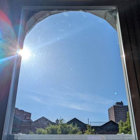 Looking through an arched window two and a half hours after sunrise the sun is above the buildings, making a dramatic lens flare with  multi-colored petals on the upper left. The sky is light blue with a few insubstantial white clouds. Pointed roofs of Harlem brownstones are silhouetted across the street, and a taller apartment building can be seen in the distance. The green tops of two trees are on the bottom and right.