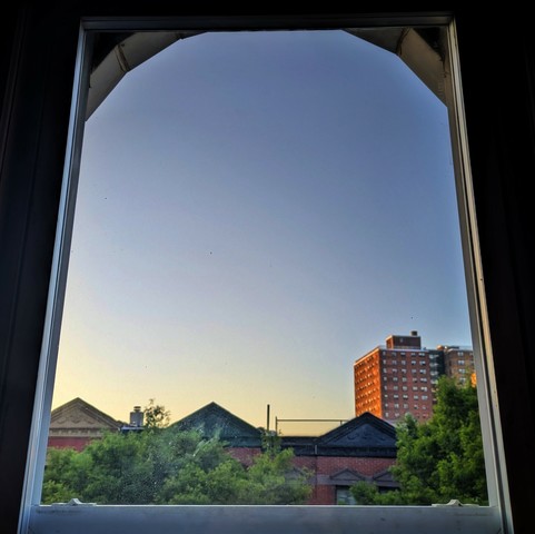 Looking through an arched window twenty seven minutes after sunrise the cloudless sky fades from light yellow at the horizon to powder blue up above. Pointed roofs of Harlem brownstones with red brickwork are across the street, and a taller apartment building can be seen in the distance. The green tops of two trees are on the bottom and right.