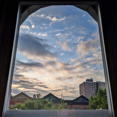 Looking through an arched window an hour after sunrise the blue sky is full of dramatic scattered grey and white clouds. Pointed roofs of Harlem brownstones with red brickwork are across the street, and a taller apartment building can be seen in the distance. The green tops of two trees are on the bottom and right.