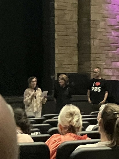 A woman addresses the audience before the film is screened. She is accompanied by another woman and the vice mayor. The event is taking place in a theater.