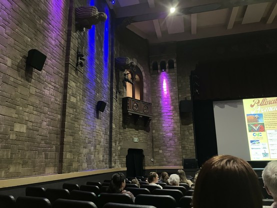 The ornate interior of the Grandin Theater featuring textured stone walls, decorative carvings, and purple lighting. Seats are filled with audience members, and a presentation screen displays event details.