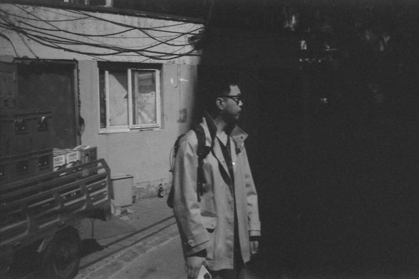Ilford FP4 @ ISO 64

English Alt Text: A person in a long coat with a backpack stands in front of a windowed building. To the left, crates and boxes are stacked on a wheeled cart, and overhead wires stretch across the scene.

中文替代文本: 一位身穿长外套、背着背包的人站在带窗户的建筑物前。左侧有一个堆满箱子的推车，上方布满电线，构成都市一角。