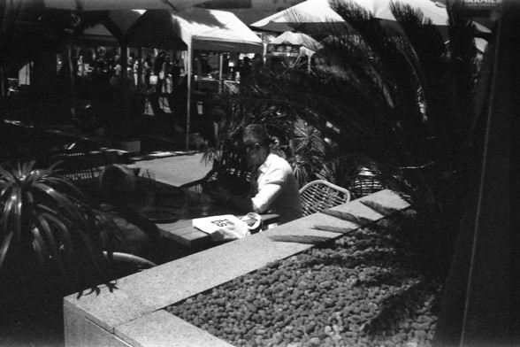 Ilford FP4 @ ISO 64

English Alt Text: A person in sunglasses sits at a table partially shaded by foliage. A bag with the letters “DS” rests on the table, and tents with people milling around suggest a lively market or outdoor event in the background.

中文替代文本: 一位戴着墨镜的人坐在桌旁，部分被植物遮住。桌上放着一个印有“DS”字样的包，背景是热闹的市场或户外活动，有帐篷和人群。