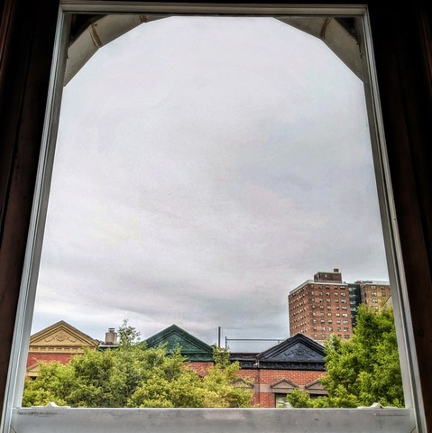 Looking through an arched window two hours after sunrise the sky is full of gray clouds. Pointed roofs of Harlem brownstones with red brickwork are across the street, and a taller apartment building can be seen in the distance. The green tops of two trees are on the bottom and right. 