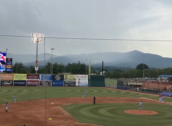A baseball game scene showing players on the field with a mountainous background. Stadium lights are on, and various sponsor banners are visible along the outfield wall. The sky appears overcast, with a curtain of rain visible beyond the outfield wall. 