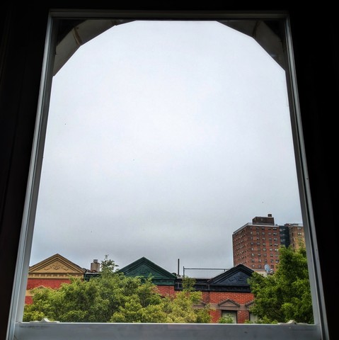 Looking through an arched window three hours after sunrise the sky is full of grey clouds. Pointed roofs of Harlem brownstones with red brickwork are across the street, and a taller apartment building can be seen in the distance. The green tops of two trees are on the bottom and right.