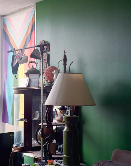 A still life featuring a whatnot crowded with small sculptures, photos, teapots, and other tchotchkes. the Progress Pride Flag is shading the window beyond and the wall is painted green. 