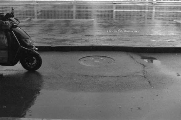 Foma FOMAPAN 400 Action (FF)

**English:**
A black and white photograph capturing a scene on a wet street. A scooter is parked on the left side of the image, and a manhole cover is visible in the center of the street. The reflection of the scooter and a person can be seen in a puddle on the wet pavement.

**Chinese:**
一张黑白照片，捕捉了湿漉漉的街道场景。一辆摩托车停在照片的左侧，一个井盖位于街道的中央。摩托车和一个人的倒影在湿漉漉的人行道上的水坑中清晰可见。
