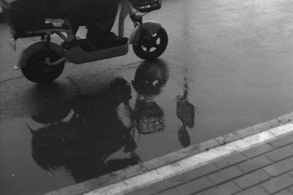 Foma FOMAPAN 400 Action (FF)

**English:**
A black and white photograph capturing a scene on a wet street. A person is seen riding a scooter on the left side of the image. The reflection of the scooter and the person is visible in a puddle on the wet pavement, creating a mirror-like effect.

**Chinese:**
一张黑白照片，捕捉了湿漉漉的街道场景。一个人骑着摩托车出现在照片的左侧。摩托车和人的倒影在湿漉漉的人行道上的水坑中清晰可见，形成镜面效果。