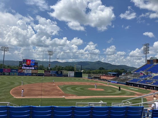 A sunny baseball field featuring players in white uniforms, with a scoreboard in the background. The scene includes green grass, a dirt infield, and mountains under a partly cloudy sky.