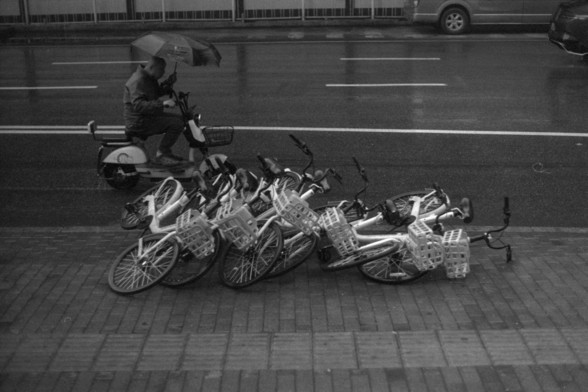 Foma FOMAPAN 400 Action (FF)

**English:**
This black-and-white photograph captures a street scene with a row of shared bicycles lying on the pavement, seemingly fallen over. A person riding a scooter with an umbrella is passing by on the left side of the image. The street is wet, indicating recent rain. In the background, there are cars on the road and a pedestrian sidewalk.

**Chinese:**
这张黑白照片拍摄了一条街道的场景，一排共享单车倒在人行道上。左侧有一位骑着带伞的踏板车的人经过。街道是湿的，表明刚刚下过雨。背景中可以看到路上的汽车和人行道。