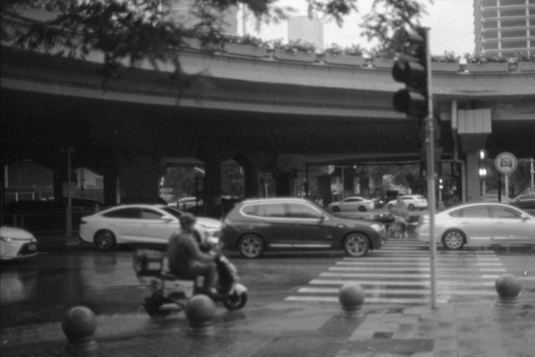 Foma FOMAPAN 400 Action (FF)

**English:**
This black-and-white image shows a busy urban intersection with several cars waiting at a traffic light. A person on a scooter is in the foreground, also waiting for the light to change. The scene is framed by an overpass or bridge above the street, and there are traffic lights and street signs visible. The street appears wet, suggesting recent rainfall.

**Chinese:**
这张黑白照片展示了一个繁忙的城市路口，几辆汽车在等待交通灯。前景中有一位骑踏板车的人也在等待信号灯变化。场景被街道上方的高架桥或立交桥所框定，可以看到交通灯和街道标志。街…
