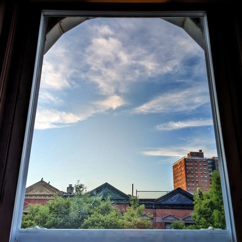 Looking through an arched window forty-five minutes after sunrise the dusty blue sky is marked with streaks and clusters of white clouds. Pointed roofs of Harlem brownstones with red brickwork are across the street, and a taller apartment building can be seen in the distance. The green tops of two trees are on the bottom and right. 