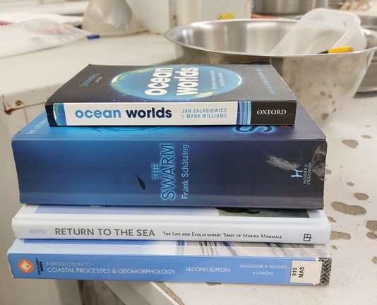 A stack of 4 blue books on my lab desk: Ocean Worlds by Jan Zakasiewicz & Mark Williams. The Swarm by Frank Schätzing, Return to the Sea by Annalisa Berta. Introduction to Coastal Processes & Geomorphology by Masselink, Hughes, & Knight.