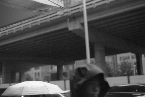 Foma FOMAPAN 400 Action (FF)

English alt text:
A black and white photo of a rainy urban scene under an overpass. A person in a hooded jacket stands in the foreground, partially visible, while a white umbrella appears in the background. Distant buildings are seen through the rain.

中文替代文字（Chinese alt text）：
一张黑白照片，展示了高架桥下雨中的城市景象。前景中部分可见一个穿着连帽夹克的人，背景中出现一把白色的伞。远处雨中依稀可见一些建筑物。