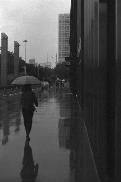 Foma FOMAPAN 400 Action (FF)

English alt text:
A black and white photo of a rainy city sidewalk. A person holding an umbrella walks away from the camera, their reflection mirrored on the wet ground. In the background, more figures with umbrellas move along the street, and tall buildings rise into the mist.

中文替代文字（Chinese alt text）：
一张黑白照片，展现城市人行道上的雨景。一个打伞的人背对镜头行走，倒影映在湿漉漉的地面上。背景中还有其他打伞的行人和高耸入云的建筑物。