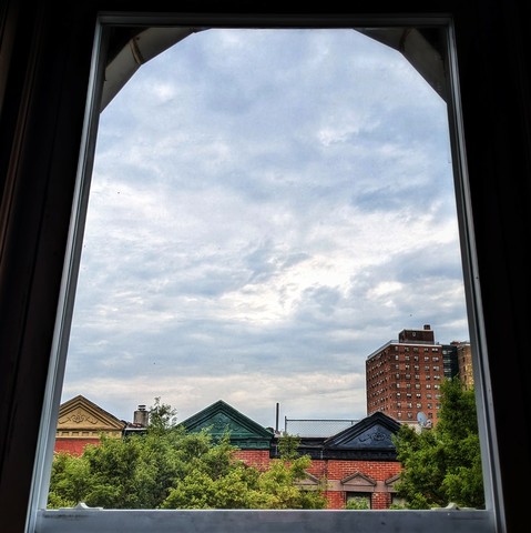 Looking through an arched window two hours after sunrise the sky is full of blue-grey clouds. Pointed roofs of Harlem brownstones with red brickwork are across the street, and a taller apartment building can be seen in the distance. The green tops of two trees are on the bottom and right.