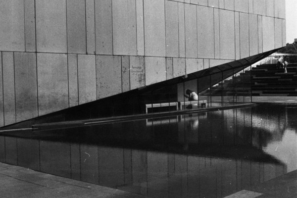 Foma FOMAPAN 400 Action (FF)

**English:**
A black and white photograph of a modern architectural structure. The image features a large, flat wall composed of vertical panels. In the foreground, there is a reflective pool of water with a slanted, angular overhang extending from the building. A person is seen. The scene captures a sense of modern design and minimalism.

**Chinese:**
一张现代建筑结构的黑白照片。图片中有一面由垂直面板组成的大型平面墙。前景是一池反光的水面，建筑物有一个倾斜的、角状的悬挑部分延伸出来。一个人正俯身。这幅画面展现了现代设计和极简主义的风格。