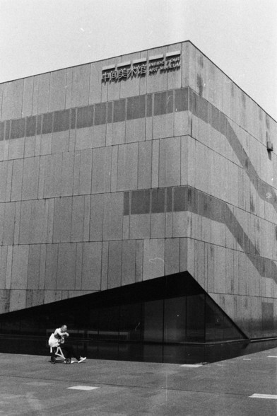 Foma FOMAPAN 400 Action (FF)

**English:**
A black and white photograph of a large, modern building with a textured facade made up of rectangular panels. The building has a sign near the top that reads "Inside-Out Art Museum." In the foreground, a person on a wheelchair. The image captures a moment of everyday life in an urban setting, juxtaposed with the imposing structure of the museum.

**Chinese:**
一张大型现代建筑的黑白照片，建筑外墙由矩形面板构成，具有纹理感。建筑顶部附近有一个标志，写着“Inside-Out艺术博物馆”。前景中，一个坐着轮椅的人。这幅图片捕捉了城市环境中日常生活…