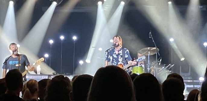A live performance featuring three musicians on stage. One vocalist is passionately singing and playing guitar, while another musician plays bass. The background displays stage lights illuminating the scene, and an audience can be seen in the foreground. Off camera, a guitar player is playing. 