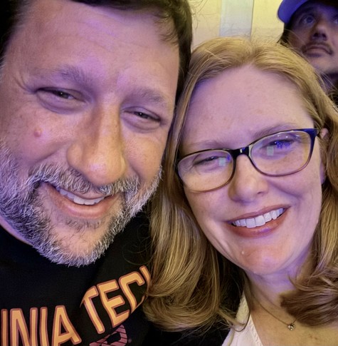 A smiling man with a beard and a woman with glasses pose closely together for a selfie. The background shows dim lighting, with another person partially visible behind them.