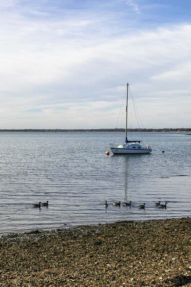A portrait view looking across very calm water from a few metres back on a shallow beach of very small shingle. There is hazy cloud covering much of the sky but warm blue shows through at the top. On the horizon there is low land stretching across. Eight birds swim in the shallows of the water just off the beach area, all passing from left to right. Anchored a dozen or more metres back from that is a small sailboat with the sail on its single tall mast furled up and covered just above the low cabin that takes up most of the vessel's top. A couple of buoys are in front and behind the boat. A reflection of the mast stretches across the water towards us, not quite reaching the shoreline against the shingle.