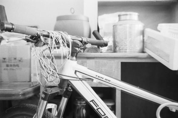 Fujifilm NEOPAN SS (FF)

**English:**
A black and white photograph of a bicycle parked indoors. The bicycle is leaning against a counter or shelf that holds various containers and jars. The handlebars of the bicycle have cables attached, and the frame has some text on it. The setting appears to be a storage area or utility room.

**Chinese:**
一张黑白室内照片，展示了一辆停放的自行车。自行车靠在一个放有各种容器和罐子的柜台或架子上。自行车的车把上有电缆连接，车架上有一些文字。场景看起来像是储藏室或杂物间。