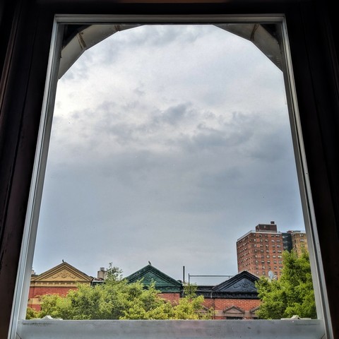 Looking through an arched window two hours after sunrise the sky is full of grey clouds. Pointed roofs of Harlem brownstones with red brickwork are across the street, and a taller apartment building can be seen in the distance. The green tops of two trees are on the bottom and right.