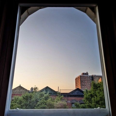 Looking through an arched window half an hour after sunrise the cloudless sky fades from light peach at the horizon to dusty blue up above. Pointed roofs of Harlem brownstones with red brickwork are across the street, and a taller apartment building can be seen in the distance. The green tops of two trees are on the bottom and right.