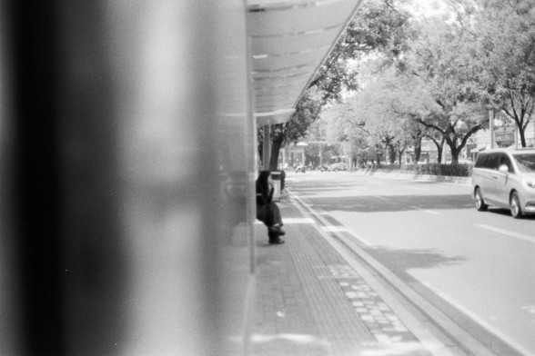 Fujifilm Neopan 100 SS (FF)

🇬🇧 English Alt Text

A black and white photo depicting a quiet street scene. A person sits alone inside a bus stop shelter on the left, partially obscured by blurriness. To the right, a car moves along a road bordered by trees. The background features buildings softened by distance, evoking a calm, introspective atmosphere.

🇨🇳 中文替代文字

一张黑白街景照片，左侧是一个公交站亭，一人独坐于长椅上，图像部分模糊。右侧道路上有一辆汽车驶过，沿途树木林立。远景中的建筑模糊朦胧，整个画面呈现出一种宁静、沉思的氛围。