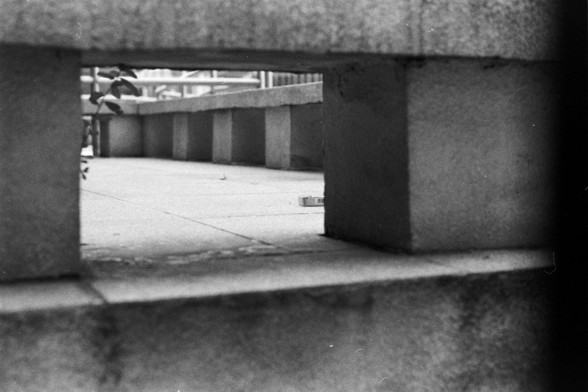 Fujifilm Neopan 100 SS (FF)

🇬🇧 English Alt Text

A black and white photograph viewed from a low angle through a rectangular gap in a concrete structure. Beyond the opening lies a paved surface with multiple concrete supports extending into the background. A small plant with broad leaves emerges on the left, adding a touch of nature to the urban geometry. In the distance, a rectangular object rests on the ground, possibly a box or piece of debris. The composition highlights tension between the …