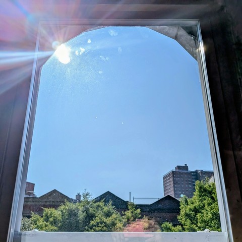 Looking through an arched window three hours after sunrise the sun is above the buildings, making a dramatic lens flare with multi-colored petals on the upper left. The sky is light blue and cloudless. Pointed roofs of Harlem brownstones are silhouetted across the street, and a taller apartment building can be seen in the distance. The green tops of two trees are on the bottom and right.