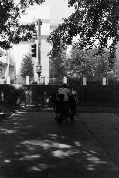Fujifilm Neopan 100 SS (FF)

---

🇬🇧 English Alt Text:
A person riding a motorcycle along a shaded urban pathway, approaching a traffic light. The light stands tall in the background with buildings and trees framing the city scene. Sunlight filters through, creating a stark contrast between the shadowed road and the bright surroundings.

🇨🇳 中文替代文字：
一位骑摩托车的人行驶在城市中阴影覆盖的小路上，正驶向远处的交通信号灯。背景中有树木和建筑，阳光透过树叶洒落在路面上，形成明暗对比，呈现出城市的日常景象。

---